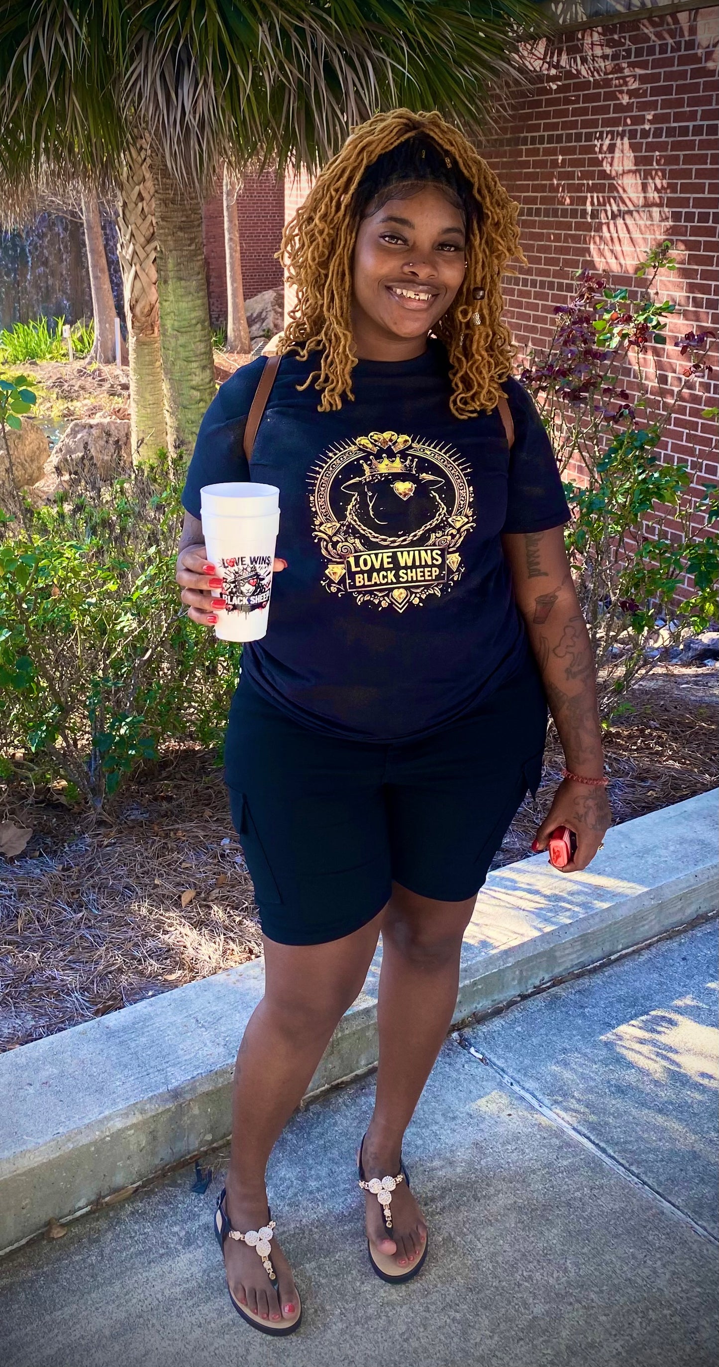 Woman holding a cup outdoors with a brick building and greenery in the background