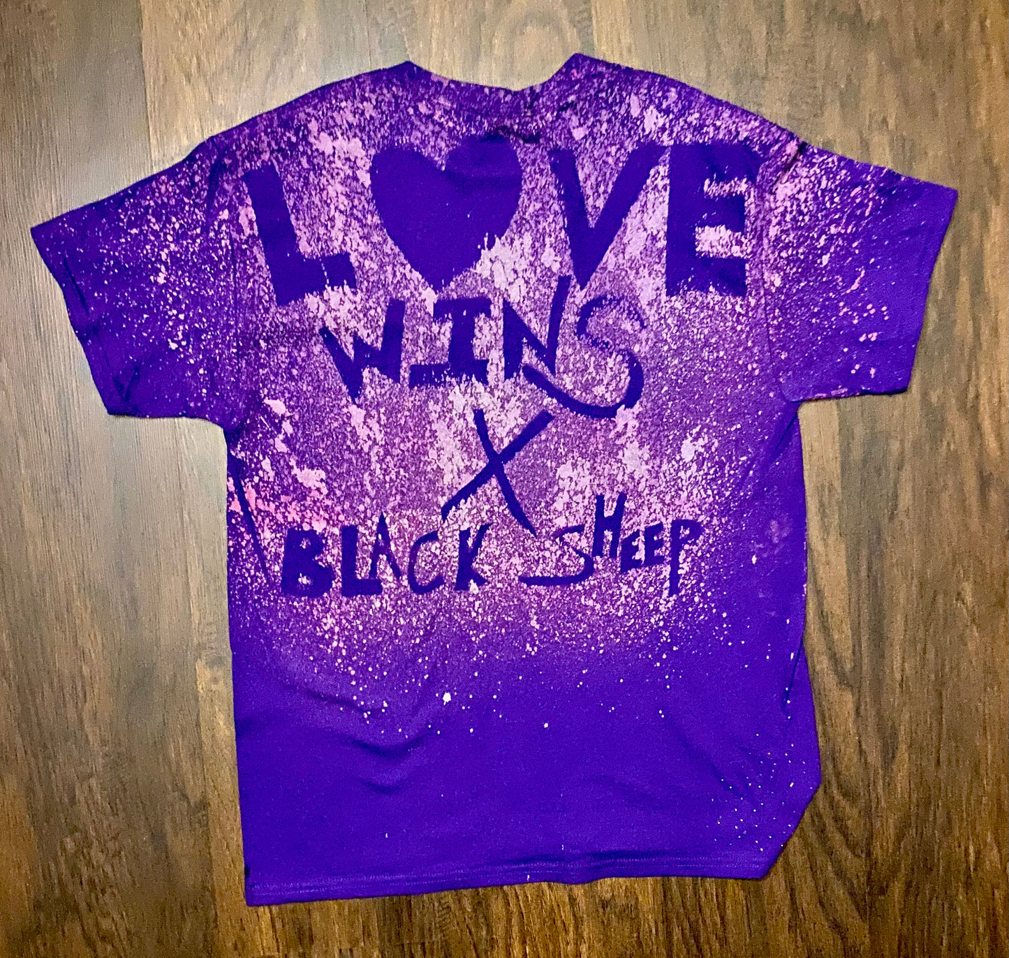 Purple t-shirt with 'LOVE WINS X BLACK SHEEP' text on a wooden surface